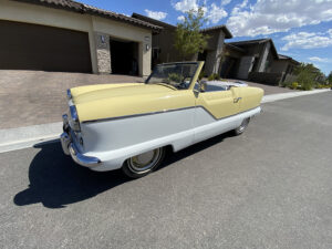 Vintage two-tone convertible (yellow top, white body) parked on a suburban street in front of modern houses with a blue sky above.