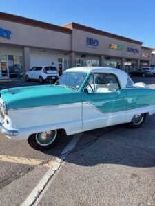 Turquoise and white vintage convertible car parked in front of a shopping plaza with storefronts visible.