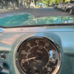 Close-up of a vintage car dashboard speedometer (0–80 mph) with a street and parked cars visible through the windshield.”, “Interior of a classic car, the mph speedometer centers the dial with fuel gauge beneath, street scene ahead.”, “Classic car dashboard speedometer on an mph scale, with a tree-lined street visible outside.”