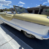 Cream and white vintage convertible parked on a sunny suburban street with houses in the background.