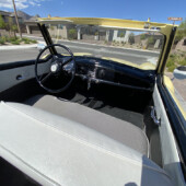 Open-top vintage convertible interior with a black dashboard, large steering wheel, and gray seats; suburb street visible through the windshield on a sunny day.