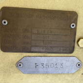 Bronze identification plate on a yellow surface reading AMERICAN MOTORS CORP with fields for BODY 34875, MODEL 561, TRIM T600, PAINT P909-P910 and two screws visible.
