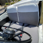 Open-top vintage car interior with gray fabric bench seat, two-seat layout, and lap belts; beige exterior visible at the door.