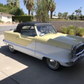 Yellow vintage convertible with black top parked in a desert-style suburban driveway with palm trees nearby.